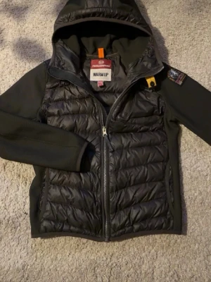 Mörkgrön cardigan från Parajumpers - Snygg mörkgrön cardigan från Parajumpers med huva och dragkedja framtill. Jackan har quiltad front och rygg i glansigt material, samt släta ärmar och sidor i stretchigt tyg. Den är äkta och de går att kollas med QR koden, helt ny, utseende mässigt ochså, den har ett litet hål som man knappast märker av, storlek 10 i barn, skriv för mer info