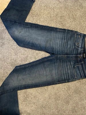 Mörkblå jeans från Jack & Jones  - Säljer ett par mörkblå jeans från Jack & Jones med klassisk femficksmodell och raka ben. Jeansen har snygga slitningar och tvättade detaljer framtill och baktill. Tillverkade i bomull med en skön passform som funkar till många olika stilar.
