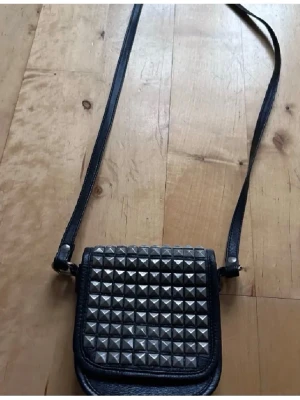 Svart axelväska med nitar - Snygg svart axelväska i läderimitation med silverfärgade nitar på locket. Väskan har justerbar axelrem och en kompakt, fyrkantig form som ger en cool och edgy vibe. Perfekt för dig som gillar detaljer som sticker ut.