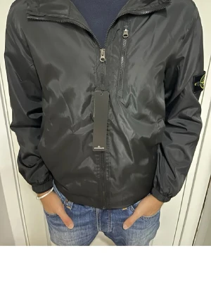 Svart windbreaker från Stone Island - Svart vindjacka från Stone Island med dragkedja framtill, bröstficka med dragkedja och klassisk logotyp-patch på vänster ärm. Jackan har elastiska muddar och är tillverkad i ett glansigt, lätt material som passar perfekt till streetwear.