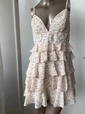 Blommig volangklänning - Söt kort klänning med flera volanger och smala axelband. Klänningen är vit med små rosa och gröna blommor, har djup urringning och dragkedja bak. Perfekt för sommaren och ger en romantisk vibe. Perfekt till midsommar 💓