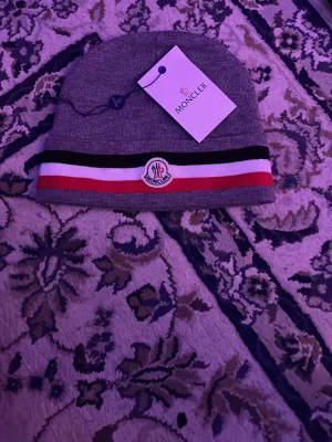 Grå Moncler mössa med logga - Snygg grå mössa från Moncler med klassisk logga framtill. Mössan har en ribbad kant med svarta, röda och vita ränder som ger en sportig look. Perfekt för dig som vill ha både stil och värme. Materialet är mjukt och stretchigt, vilket gör den bekväm att bära.