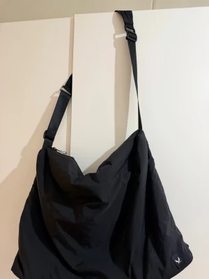 Stor svart axelväska i nylon - En oversized svart axelväska i mjukt nylonmaterial med justerbar axelrem och enkel design. Väskan har en diskret logga i hörnet och passar perfekt för dig som vill ha plats för allt. Snygg och praktisk till vardagslooken.