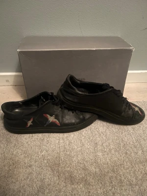 Svarta sneakers från Axel Arigato - Säljer ett par svarta sneakers från Axel Arigato med broderade fåglar i färgerna röd, blå och vit på sidan. Skorna har en klassisk låg profil, svarta snören och är tillverkade i läder. Perfekta för dig som vill ha en stilren men unik look. är ganska slitna som man ser på bilderna. Storlek 44