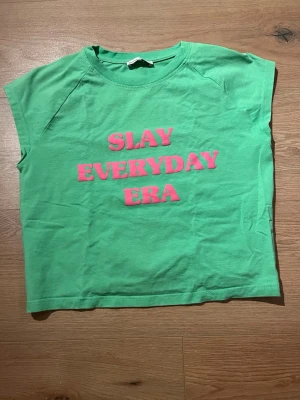 Grön topp med rosa text BERSHKA - Grön topp i bomull från BERSHKA med trycket 'SLAY EVERYDAY ERA' i rosa framtill. Modellen är ärmlös med rund hals och har en avslappnad passform. Perfekt för dig som vill sticka ut med färg och budskap.