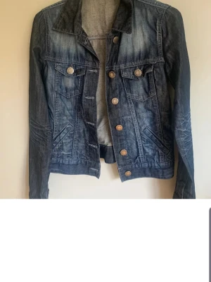 Mörkblå klassisk jeansjacka - Snygg mörkblå jeansjacka med slitningar och tvättade detaljer. Jackan har två bröstfickor med knappar, två sidofickor och metallknappar framtill. Figurnära passform och klassisk krage ger en tidlös look. Perfekt att slänga över en hoodie eller t-shirt.