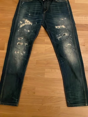Mörkblå slitna jeans med raka ben - Säljer ett par mörkblå jeans med tydliga slitningar och hål på framsidan av benen. Jeansen har raka ben och klassisk femficksdesign. Perfekta för en avslappnad streetstil.