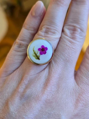 Guldfärgad ring med en blomma - Snygg guldfärgad ring med justerbar öppning och en rosa blomma och grön mossa.. Perfekt accessoar för dig som gillar unika och färgglada smycken.