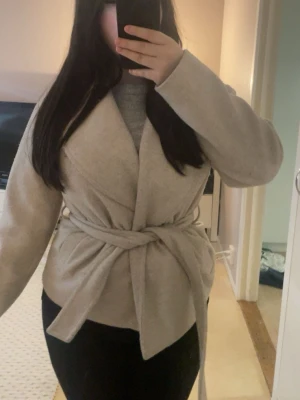 Beige kappa  - Snygg beige kappa med bred krage och knytskärp i midjan. Kappan har en mjuk och stilren design, perfekt för våren och för att hålla värmen under kyliga dagar. Materialet ser ut att vara ullblandning och kappan har en avslappnad passform.