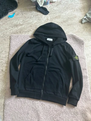 Svart hoodie från Stone Island - Svart hoodie från Stone Island med dragkedja framtill och klassisk logopatch på vänster ärm. Tröjan har huva med dragsko och två fickor framtill. Tillverkad i mjukt bomullsmaterial, perfekt för en avslappnad streetwear-look. Helt ny använt 1 gång. 