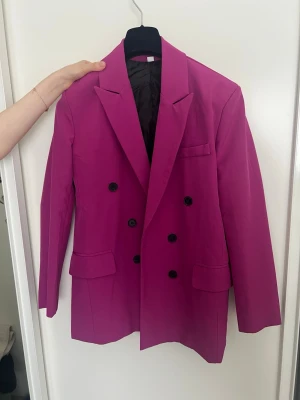 Fuchsia dubbelknäppt kavaj - Stilren fuchsia kavaj med dubbelknäppning och svarta knappar. Klassisk krage, två fickor med lock och en bröstficka. Fodrad insida och rak passform. Perfekt för dig som vill sticka ut med färg. Materialet känns slätt och håller formen snyggt.
