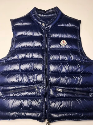 Mörkblå dunväst från Moncler - Snygg mörkblå dunväst från Moncler med glansig finish och klassisk logga på bröstet. Västen har ståkrage, två dragkedjefickor framtill och quiltad design. Perfekt för lager på lager och streetstyle.