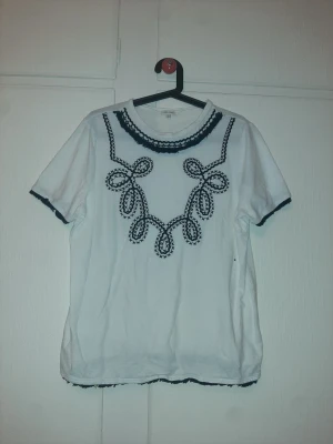 Vit t-shirt med broderi River Island - Vit t-shirt från River Island med svart broderat mönster framtill och runt kragen. Klassisk passform och korta ärmar. Tillverkad i mjuk bomull som känns skön mot huden. Snygga detaljer med broderi som ger en unik look.