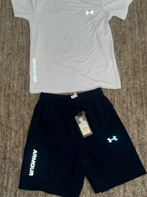 Under Armour träningsset herr - Säljer ett sportigt träningsset från Under Armour med en vit t-shirt och svarta shorts. T-shirten har reflekterande logga och textdetaljer, rund hals och korta ärmar. Shortsen har resår i midjan, reflekterande logga och text längs benet. Perfekt för gym eller löpning.
