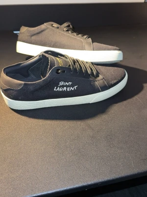 Svarta Saint Laurent sneakers - Snygga svarta sneakers från Saint Laurent med vit sula och handskriven logga på sidan. Skorna har klassisk låg profil, svarta skosnören och är tillverkade i canvas med detaljer i läder. Perfekta för en stilren och trendig look.
