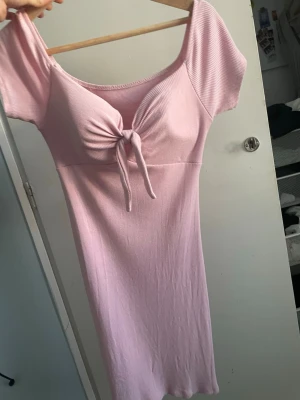 Ljusrosa ribbad klänning med knyt - Söt ljusrosa klänning i ribbat material med korta ärmar och knyt framtill vid bysten. Klänningen har en kroppsnära passform och en mjuk, stretchig känsla. Perfekt för en trendig och avslappnad look.