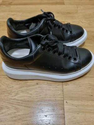 Svarta Alexander McQueen sneakers - Stilrena svarta sneakers från Alexander McQueen med chunky vit sula och svarta skosnören.  Perfekta för dig som gillar en clean och modern look.
