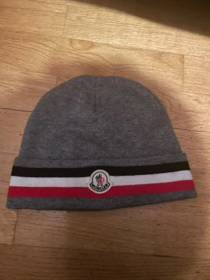Grå Moncler mössa med ränder - Snygg grå mössa från Moncler med klassiska svarta, vita och röda ränder runt kanten. Mössan har Moncler-loggan framtill och är tillverkad i mjukt stickat material som känns skönt mot huvudet. Perfekt accessoar för kyliga dagar.