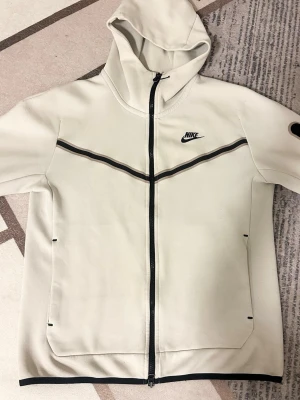 Vit Nike tech fleece  - Snygg vit hoodie från Nike med hel dragkedja och svarta detaljer. Hoodien har en huva, två stora fickor framtill och en svart Nike-logga på bröstet. Materialet är mjukt och bekvämt, perfekt för en avslappnad streetwear-look.  Storlek S men passar som M också priset går att diskuteras 
