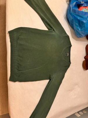 Stickad tröja - Herrmodell, grön stickad tröja. Den är lagad i armhålorna. Detta syns dock knappt när man har på sig den. Finns ett mindre hål på armen men det går laga.