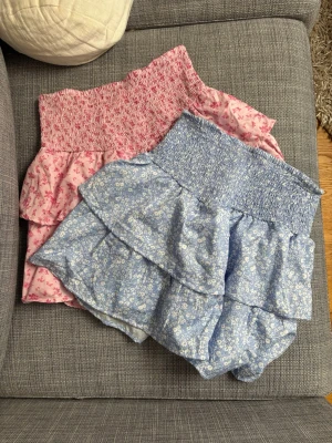 Blommiga volangkjolar i rosa och blått - Två korta kjolar med smockad midja och dubbla volanger. En är ljusblå med små vita blommor och en är ljusrosa med rosa blommor. Kjolarna är gjorda i ett tunt, mjukt bomullstyg som känns perfekt för varma dagar. Kontakta mig om du vill köpa en kjol separat, annars säljs kjolarna ihop 😊