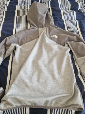 Grå hoodie från The North Face - Snygg hoodie från The North Face i ljusgrått med mörkgrå detaljer på ärmar, axlar och sidor. Dragkedja framtill och justerbar huva. Tillverkad i mjukt och bekvämt material, perfekt för chill dagar eller när du vill ha en sportig look. Logga på bröstet.