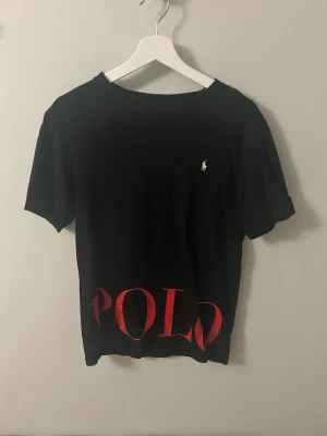 Svart t-shirt från Polo Ralph Lauren - Snygg svart t-shirt från Polo Ralph Lauren med röd POLO-logga nertill och klassisk liten vit logga på bröstet. T-shirten har rund hals och korta ärmar, perfekt för en stilren och avslappnad look.