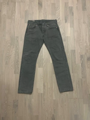 Levi’s 501 Grå - Storlek W31 L32 - Säljer ett par grå jeans från Levi's med klassisk femficksmodell och raka ben. Jeansen har metallknappar och nitar samt den ikoniska läderlappen bak i midjan. Perfekta för en avslappnad och stilren look.