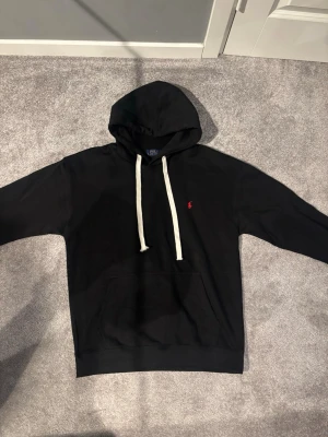 Ralph Lauren hoodie - Säljer en svart hoodie från Polo Ralph Lauren med vit dragsko och klassisk röd logga på bröstet. Hoodien har känguruficka framtill och ribbade muddar vid ärmar och nederkant. Perfekt för en avslappnad och stilren look.