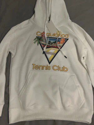 Vit Casablanca Tennis Club hoodie - Snygg vit hoodie från Casablanca med färgglad grafik och texten 'Tennis Club' på bröstet. Hoodien har en stor magficka, dragsko i huvan och tryck med palmer och byggnader i retrostil. Perfekt för dig som gillar streetwear och sportig vibe.
