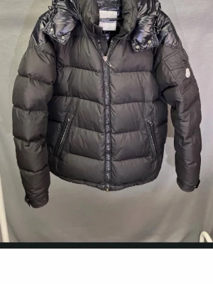 Svart dunjacka från Moncler - Snygg svart dunjacka från Moncler med hög krage och avtagbar huva. Jackan har två dragkedjeförsedda fickor fram och en Moncler-logga på ärmen. Skick 5/10 flera defekter förekommer, cartoontag lossnat etc. Klassisk puffer-modell med quiltade paneler och blanka detaljer vid axlarna.