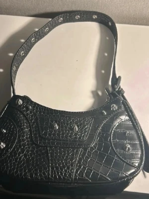 Svart axelväska i krokopräglat material - Snygg svart axelväska i krokopräglat konstläder med silverfärgade nitar och detaljer. Väskan har en bred axelrem med metallhål och dragkedja baktill. Perfekt för dig som gillar edgy och trendig stil.