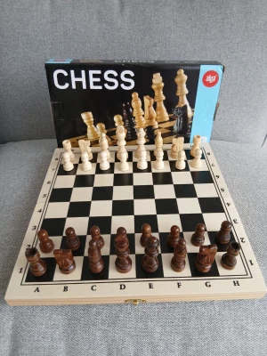 Schackspel i trä från Alga - Säljer ett klassiskt schackspel från Alga med schackbräde och pjäser i trä. Brädet är svartvitt med bokstäver och siffror längs kanterna och fungerar även som förvaringslåda. Pjäserna är i två färger, mörkbruna och ljusa, och spelet levereras med instruktioner.