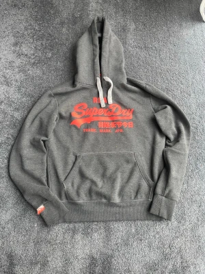  Superdry hoodie - Snygg grå hoodie från Superdry med stor röd logga och japansk text på bröstet. Klassisk känguruficka framtill, ribbade muddar och vit dragsko i huvan. Perfekt för en avslappnad och trendig stil. Tillverkad i mjukt bomullsmaterial.