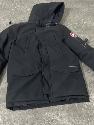 Svart parkas från Canada Goose - Svart parkasjacka från Canada Goose med huva, dragkedja och tryckknappar. Jackan har flera fickor framtill och en patch med logga på ärmen. Klassisk, rak modell i slitstarkt material som passar perfekt för kalla dagar.