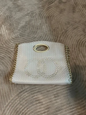 Beige handväska med guldkedja - Snygg beige handväska i flätad look med guldfärgade detaljer och kedja. Väskan har ovala handtag i metall och dekoration med nitar framtill i form av två cirklar. Perfekt accessoar för att lyfta din outfit. Den är knappt använd 