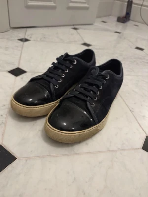 Svarta sneakers med lackad tå - Snygga svarta sneakers med blank lackad tå och ovandel i mocka. Skorna har svarta skosnören, silverfärgade öljetter och en beige sula som ger en cool kontrast. Perfekta för dig som gillar stilrena men ändå unika detaljer. Endast skorna medföljer 