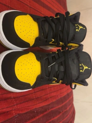 Air Jordan 1 Mid svart/gul - Snygga Air Jordan 1 Mid sneakers i svart och gult med vita detaljer. Klassisk Jumpman-logga på plösen och gul sula med Nike-märke undertill. Ovandelen är i läder och textil, med svarta skosnören och perforerad tåbox för extra ventilation.