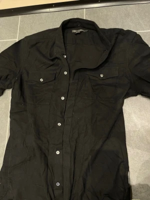 Svart skjorta från French Connection - Stilren svart skjorta från French Connection med klassisk krage och silverfärgade knappar. Skjortan har två bröstfickor med lock och knapp, samt lång ärm. Tillverkad i mjuk bomull som känns skön mot huden.