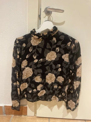 Blommig svart blus med volangkrage - Snygg svart blus med beige blommönster och volangdetaljer vid kragen. Blusen har långa ärmar och är tillverkad i ett tunt, lätt material som ger en luftig känsla. Perfekt för dig som gillar romantiska och trendiga plagg med unik touch.