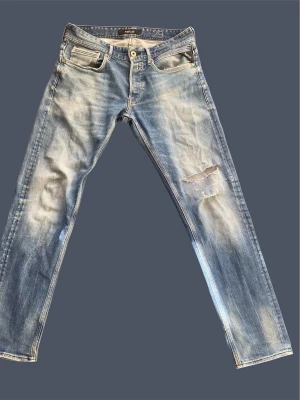 Schysta Replay jeans  - Säljer ett par grischiga blå jeans från Replay  med slitningar och hål på ena benet. Jeansen har klassisk femficksmodell, raka ben och ljusa tvättade partier för en trendig look. Perfekta för dig som gillar en avslappnad och cool stil.