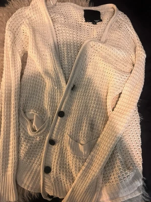 Vit stickad kofta från 157 - Snygg vit stickad kofta från 157 med knappar framtill och två fickor. Koftan har ett luftigt mönster och ribbade muddar vid ärmslut och nederkant. Perfekt att slänga på sig när det är lite kyligt ute.