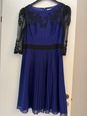 Blå klänning med spets från Karen Millen - Elegant blå klänning från Karen Millen med svarta spetsdetaljer och broderier på axlar och ärmar. Klänningen har halvtransparenta trekvartsärmar, markerad midja och plisserad kjol. Perfekt för dig som vill ha en unik och stilren look.
