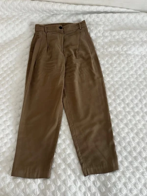 H&M bruna byxor - Byxor med veck framtill och knappstängning. De har en avslappnad passform och är lämpliga för olika tillfällen.