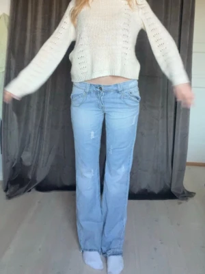 Ljusblå bootcut jeans med slitningar - Säljer ett par ljusblå bootcut jeans med slitna detaljer och dragkedjor på bakfickorna. Jeansen har låg midja och klassisk femficksdesign. Perfekta för en avslappnad och trendig look. Coola slitningar framtill på byxorna