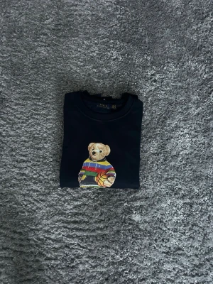 Marinblå Ralph lauren tröja  - Cool marinblå sweatshirt från Polo Ralph Lauren med ikoniskt björnmotiv på bröstet. Björnen har en färgglad randig tröja och håller en basketboll. Tröjan har rund hals och ribbade muddar vid ärmar och midja. Perfekt för en avslappnad streetwear-look.