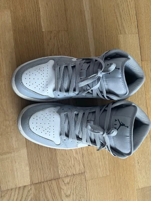 Gråa Air Jordan 1 Mid sneakers - Snygga Air Jordan 1 Mid sneakers i grått, vitt och svart. Skorna har klassisk siluett med perforerad tå, gråa skosnören och ikoniska Nike swoosh på sidan. Tillverkade i läder och textil för en clean och sportig look.