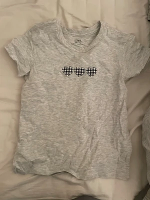 Grå t-shirt med hjärtan från Cotton On - Ljusgrå t-shirt från Cotton On med tre rutiga hjärtan broderade på bröstet. Klassisk rund hals och korta ärmar. Tillverkad i mjuk bomull, perfekt för en avslappnad och trendig stil.