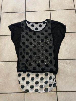 Svart mesh topp med prickigt mönster storlek S/M - Snygg svart mesh topp med stora svarta prickar och transparenta partier. Toppen har en lös passform, rund halsringning och dekorativa stenar framtill som formar ett mönster. Understycket är vitt med svarta prickar för en cool lager-på-lager-look.