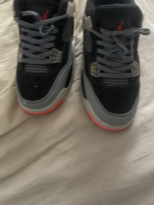 Svarta Air Jordan 4 sneakers - Säljer ett par svarta Air Jordan 4 sneakers med grå och röda detaljer. Skorna har meshpaneler på sidorna, plastdetaljer och synlig Air-sula. Gråa skosnören och tydlig Jumpman-logga på plösen. Snygg streetstil och klassisk siluett.